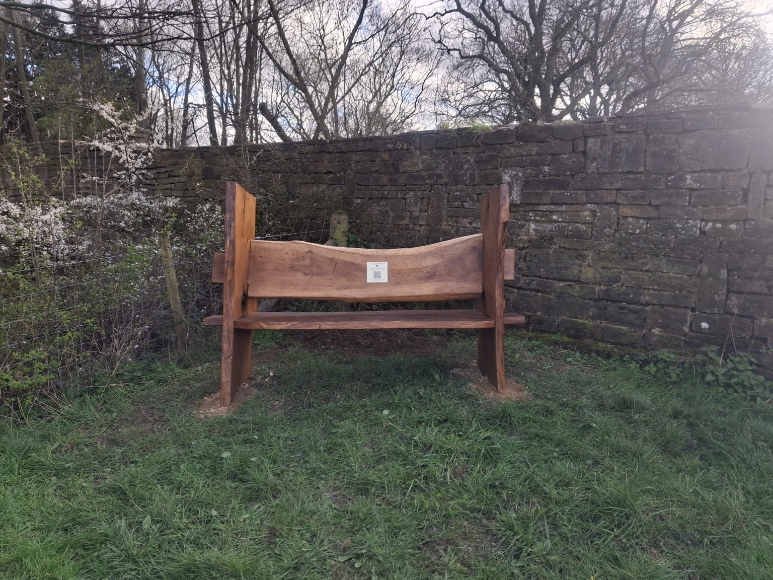 A handmade wooden memorial bench beside a dry stone wall in early spring